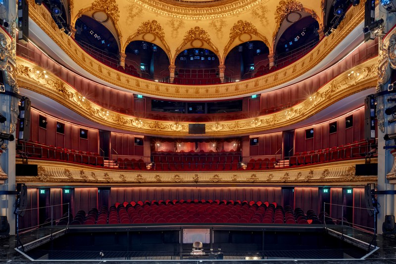 Das Bild zeigt den Saal des Stadttheaters vom der Bühne aus betrachtet. Im Kontrast zur schlichten Bestuhlung in rot stehen die goldfarbig verzierten Balkone sowie das Deckengewölbe