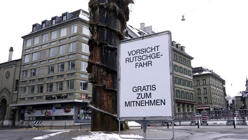 Im Hintergrund steht der Meret-Oppenheim-Brunnen, im Vordergrund ein Plakat mit der Aufschrift «Vorsicht, Rutschgefahr. Gratis zum Mitnehmen.»