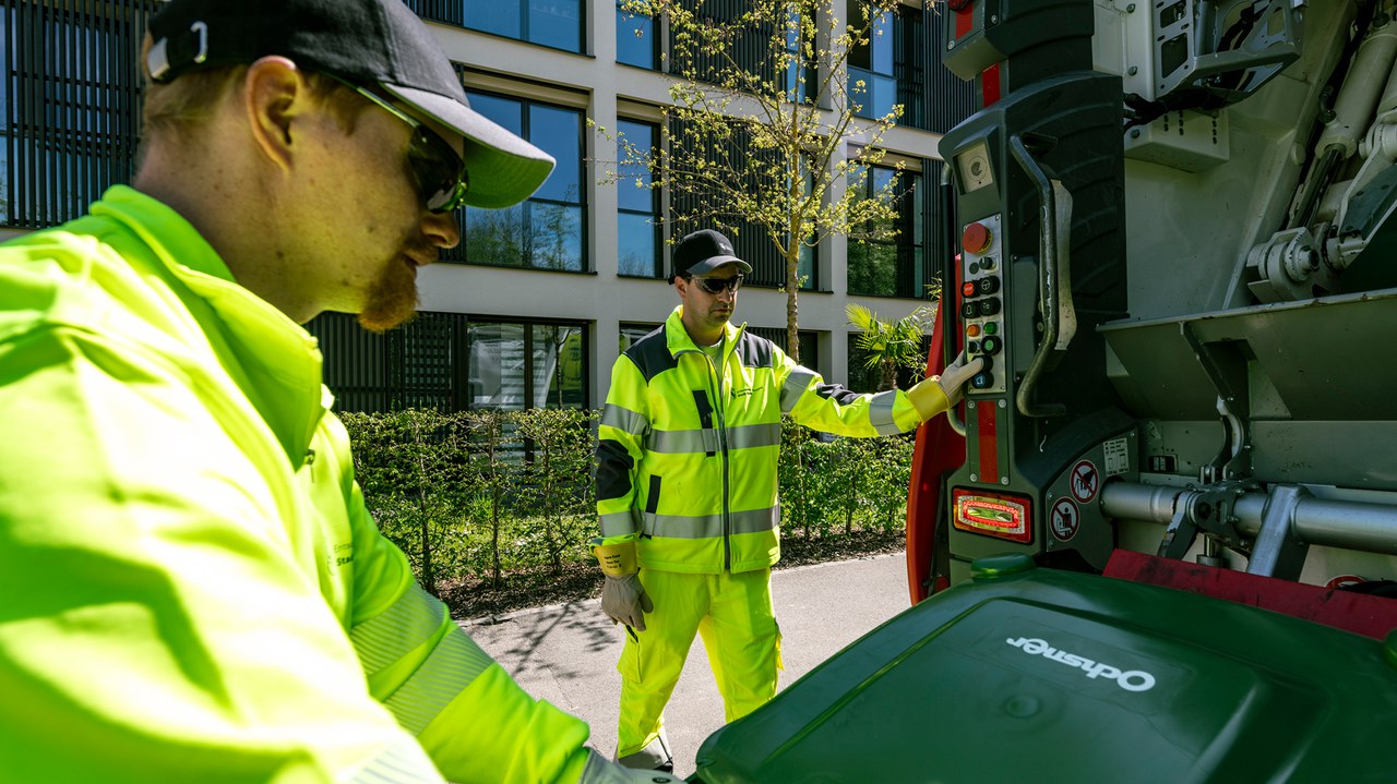 Ein Mitarbeitender hängt einen Grüngutcontainer an den Kehrichtlastwagen, ein zweiter Mitarbeiter steht an den Bedienknöpfen des Lastwagens.