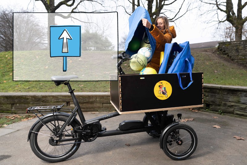 Das Bild zeigt eine Lehrperson, die Fussbälle aus einer blauen Tasche in die Ladekiste eines Cargobikes kippt. Oben links im Bild ist das Icon des Handlungsfelds 4 - Mobilität - dargestellt.